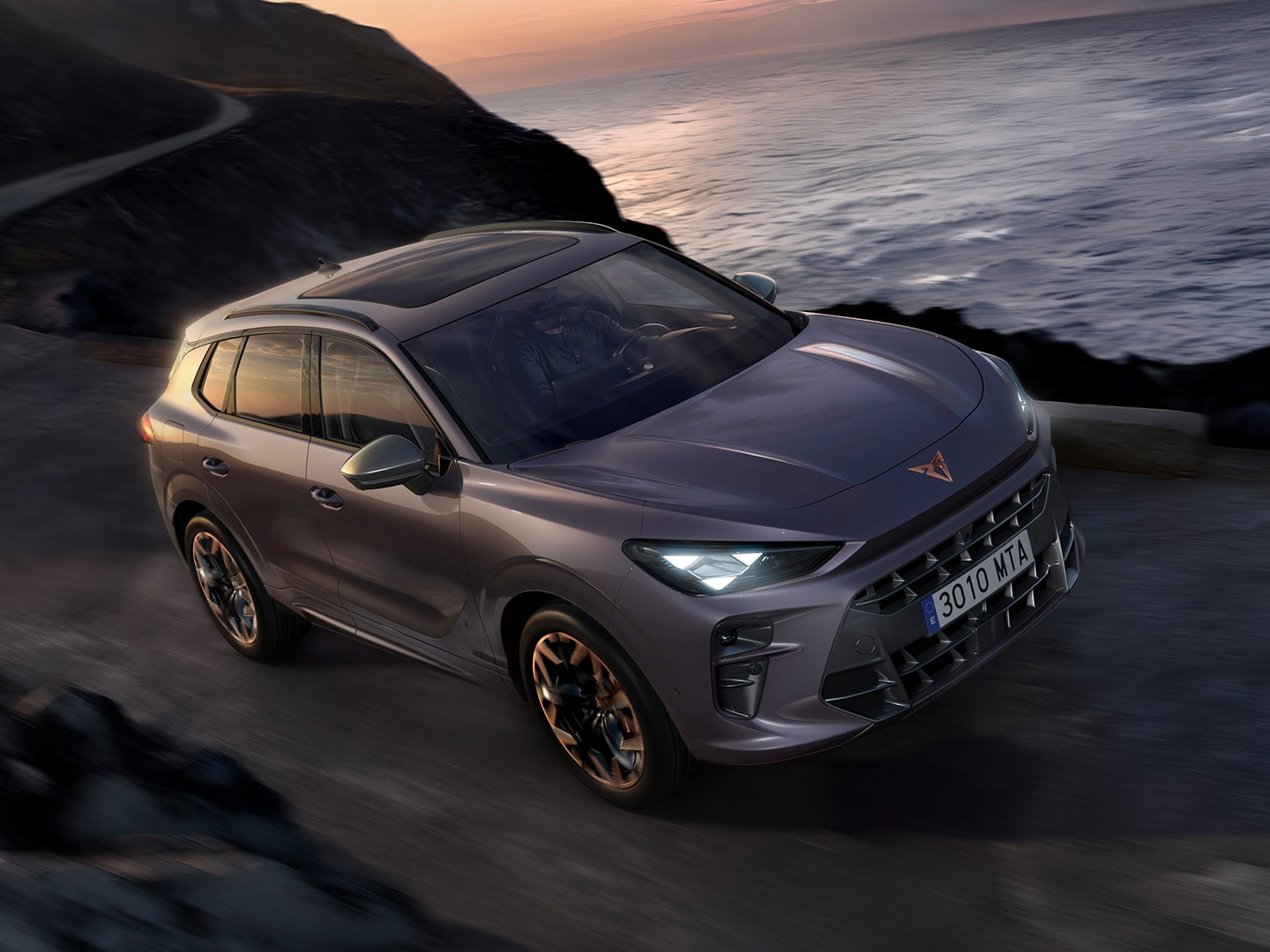 CUPRA Terramar, with dynamic chassis control (DCC), driving along a coastal road featuring dark void paint, and 20” Hadron Copper alloys. 