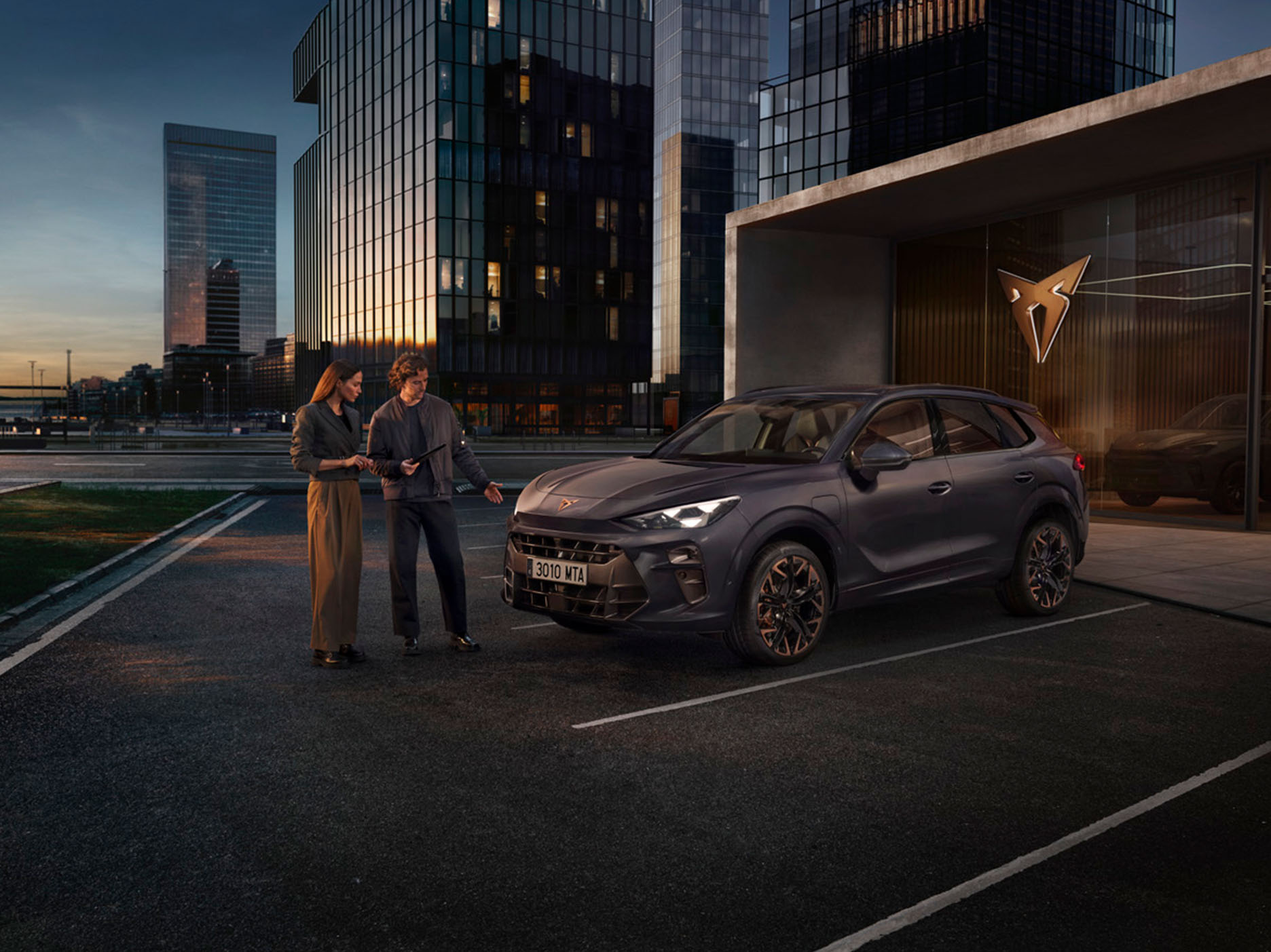 Two individuals standing in front of the CUPRA Terramar stationed in a parking next to a CUPRA building, in a city landscape.