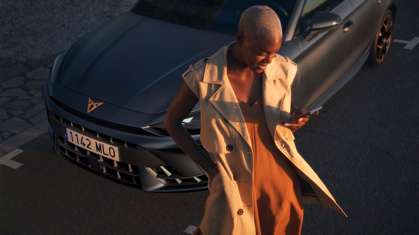 A person with short-cropped hair wearing a beige sleeveless trench coat over a rust-colored dress, looking at a smartphone and standing in front of a CUPRA car, captured in warm golden hour light.